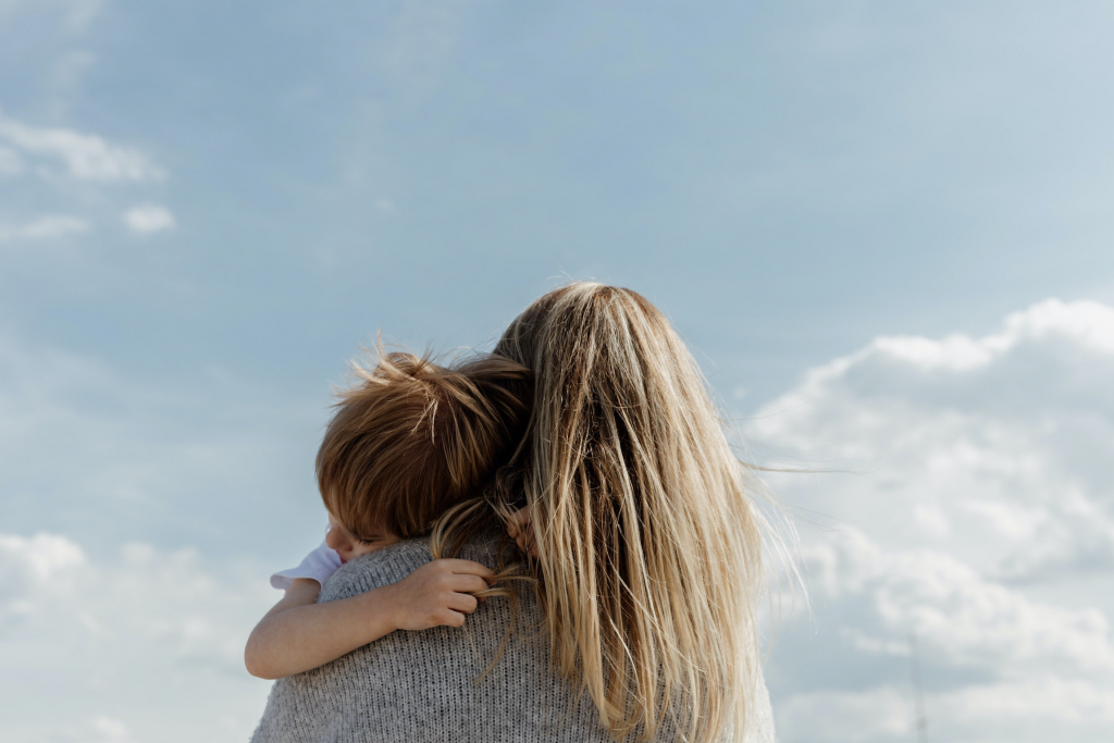 A mother holding and hugging her son. The point of view is from their behind and from the waist up with the blue sky as a background.